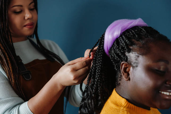 Close-up of confident hairdresser braiding hair to smiling female customer on blue background