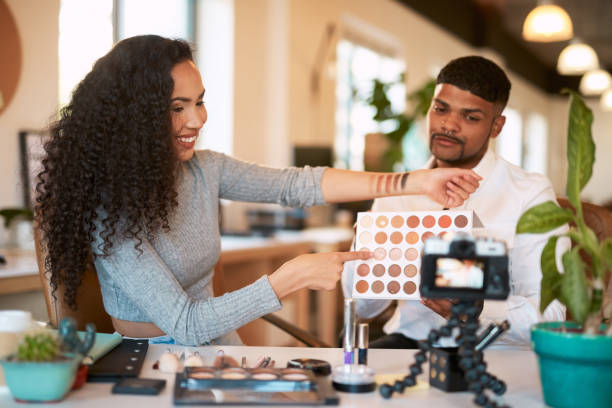 A female makeup artist holds up her arm with makeup swatches to a video camera while a male client sits beside her.