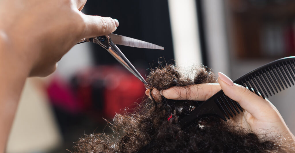 Cropped view of african american hairstylist holding comb and scissors near hair of client, banner